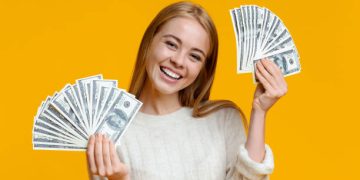 Young investor. Happy millennial girl holding lots of dollars on orange background with free space